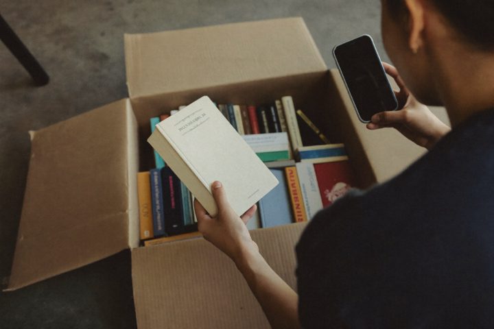 A woman crouching beside an open box of books, holding one book in one hand and her phone in the other.