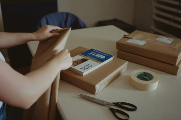 Hands wrapping a book in brown kraft paper at a home dining table, next to already-wrapped parcels.