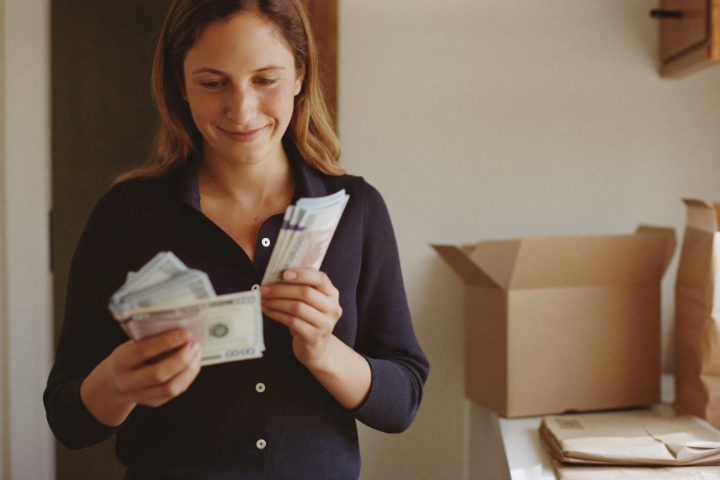A woman holding the cash from a batch of sold books, with an empty book box and wrapped parcels on the counter behind her.
